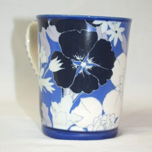 May include: A ceramic mug with a blue rim and handle, adorned with a floral pattern. The design features large navy blue flowers with white and light blue accents against a blue background. The mug is a standard size for beverages.
