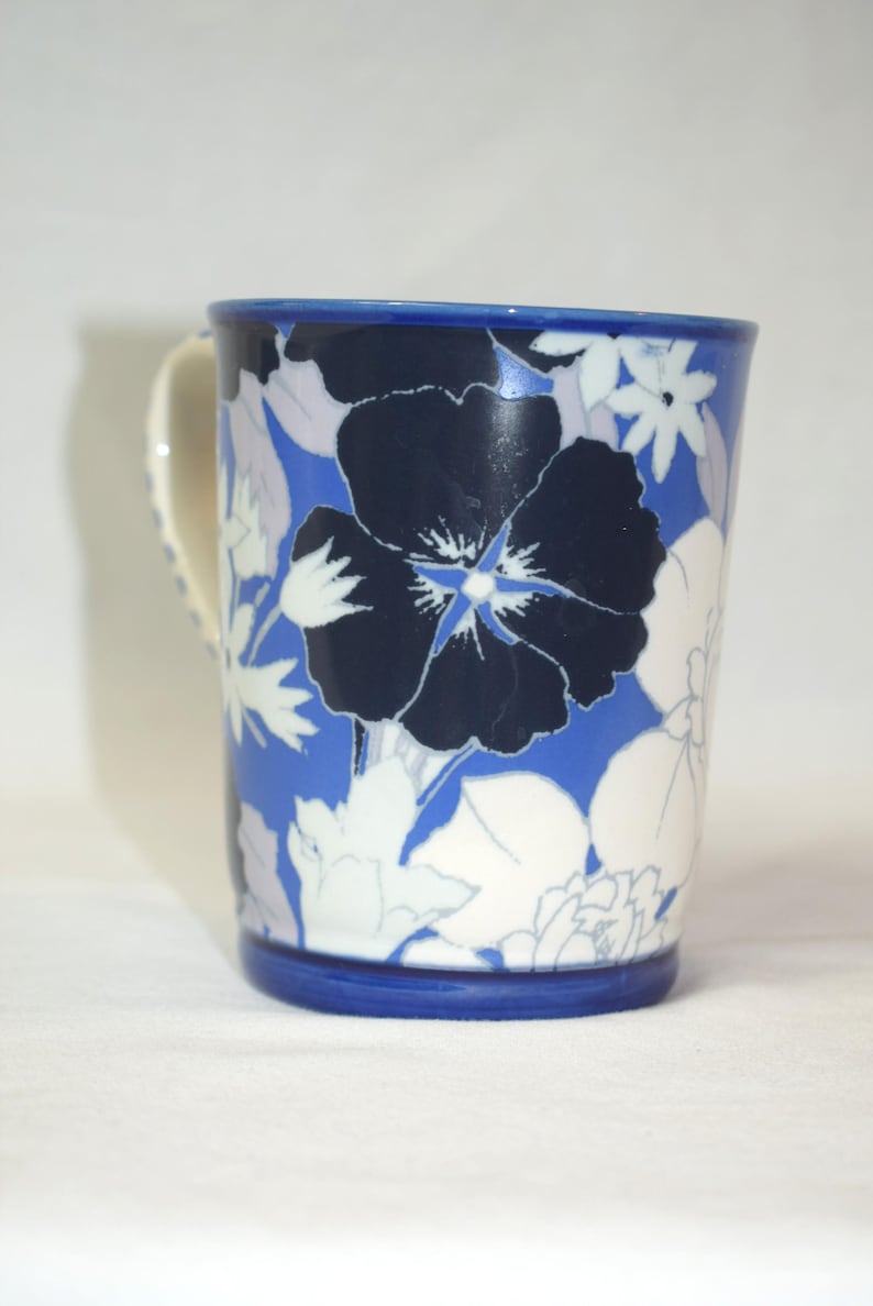 May include: A ceramic mug with a blue rim and handle, adorned with a floral pattern. The design features large navy blue flowers with white and light blue accents against a blue background. The mug is a standard size for beverages.