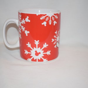 May include: Red ceramic mug with a white snowflake pattern and Mickey Mouse ears. The mug has a white handle.