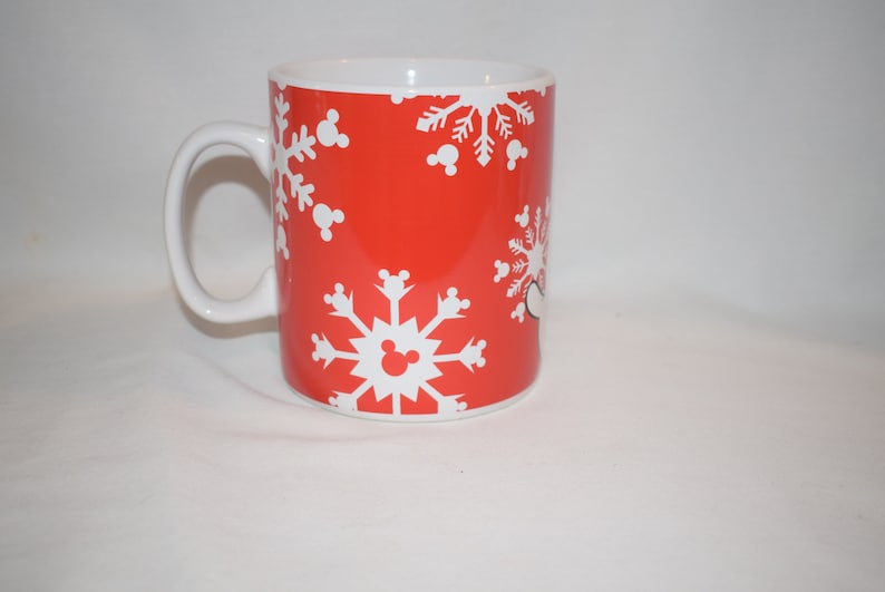 May include: Red ceramic mug with a white snowflake pattern and Mickey Mouse ears. The mug has a white handle.