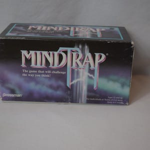 May include: A Mind Trap board game box with a black background and the game's title in a stylized font. The box features the text "The game that will challenge the way you think!" and is suitable for ages 12 and up.