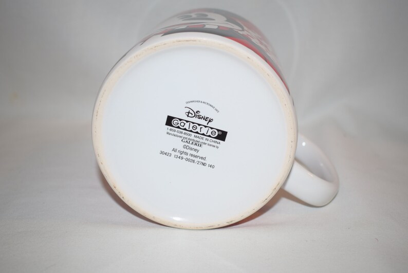 May include: White ceramic mug with a black and red Disney Galerie logo. The mug has a black handle and the text "Disney Galerie 1-800-538-4500 MADE IN CHINA Manufacturer for all items by GALERIE &copy;Disney All rights reserved. 30423 1349-0026/2710 140" printed on the bottom.