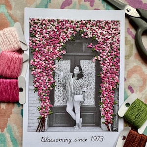 Op de afbeelding: Een zwart-witfoto van een persoon die voor een deur staat die versierd is met roze en groene geborduurde bloemen. De foto is omlijst met een witte rand en heeft de tekst "Blossoming since 1973" onderaan.