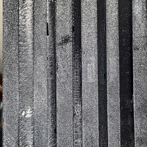 Pu&ograve; includere: Un oggetto rettangolare nero con una superficie ruvida caratterizzata da cinque scanalature verticali parallele di larghezza variabile. Il materiale appare opaco e granuloso, con distinti canali lineari nel blocco.