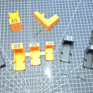 May include: A collection of 3D printed plastic guides in black, orange, and purple, featuring printed fractional labels including 3/4, 1/2, 3/8, and 1/4. These components vary in shape, with some featuring open channels and others having angled or curved profiles, all designed for precise alignment tasks.