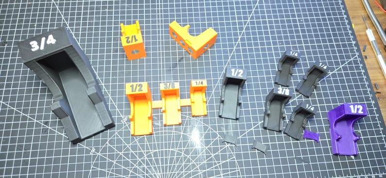 May include: A collection of 3D printed plastic guides in black, orange, and purple, featuring printed fractional labels including 3/4, 1/2, 3/8, and 1/4. These components vary in shape, with some featuring open channels and others having angled or curved profiles, all designed for precise alignment tasks.