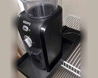 May include: A black Krups coffee grinder with a clear plastic hopper on top, a silver control dial, and a power button. The appliance features a clear collection container at the front and sits on a black rectangular tray.
