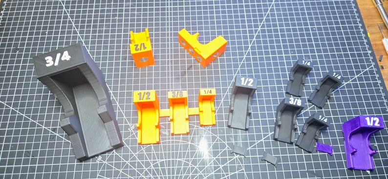 May include: A collection of 3D printed plastic corner clamping jigs in various sizes and colors, including black, orange, and purple. Each piece is clearly labeled with its specific size, such as 3/4, 1/2, 3/8, and 1/4, indicating the measurement for each tool.