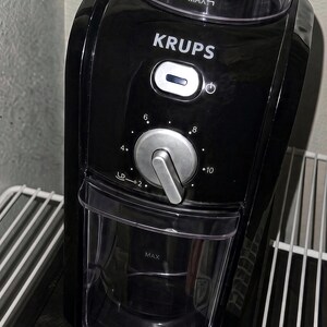 Pu&ograve; includere: Un macinacaff&egrave; nero Krups dotato di tramoggia superiore trasparente con linea di riempimento MAX, manopola centrale argentata con impostazioni da 2 a 10, pulsante di accensione e contenitore di raccolta trasparente.