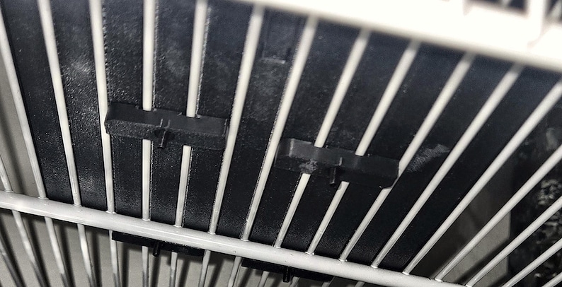 Pu&ograve; includere: Uno scaffale in filo metallico bianco con due clip in plastica nera fissate alle barre verticali. Le clip sono rettangolari con una piccola linguetta sporgente sulla parte anteriore, progettate per fissare la struttura in filo.