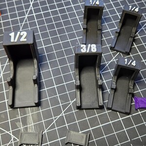 May include: A collection of five black 3D-printed plastic brackets with white embossed labels. Three brackets are marked 1/4, one is marked 3/8, and one is marked 1/2. Two small rectangular black plastic pieces are positioned at the bottom.