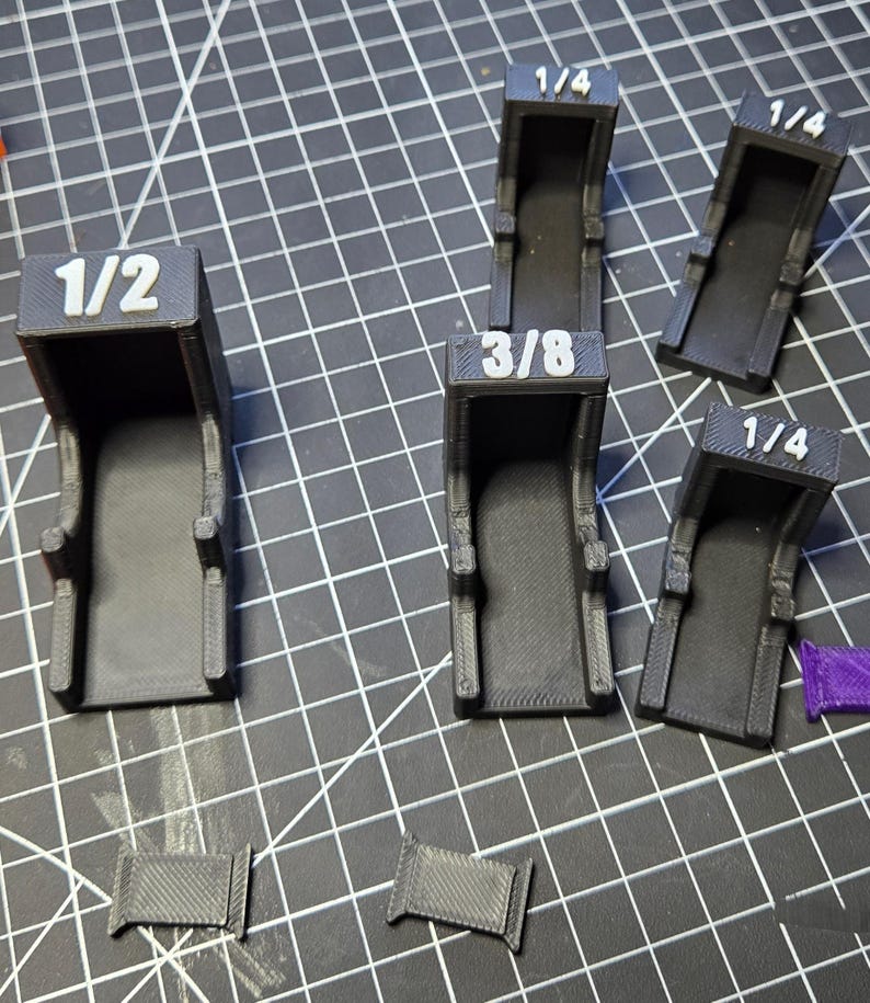 May include: A collection of five black 3D-printed plastic brackets with white embossed labels. Three brackets are marked 1/4, one is marked 3/8, and one is marked 1/2. Two small rectangular black plastic pieces are positioned at the bottom.