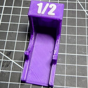 May include: A purple 3D-printed plastic bracket with a flat base and a vertical back panel. The top of the back panel features the white text 1/2 embossed on the surface. The object has a textured finish consistent with additive manufacturing.