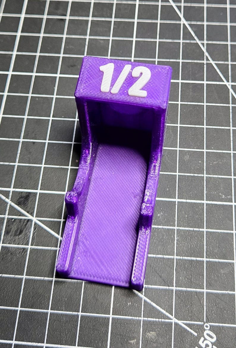 May include: A purple 3D-printed plastic bracket with a flat base and a vertical back panel. The top of the back panel features the white text 1/2 embossed on the surface. The object has a textured finish consistent with additive manufacturing.
