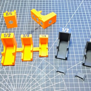 May include: A collection of 3D printed L-shaped corner clamps in black, orange, and purple. Each piece features white printed fractions including 3/4, 1/2, 3/8, and 1/4. The set includes various sizes of brackets and small rectangular inserts.