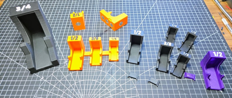 May include: A collection of 3D printed L-shaped corner clamps in black, orange, and purple. Each piece features white printed fractions including 3/4, 1/2, 3/8, and 1/4. The set includes various sizes of brackets and small rectangular inserts.