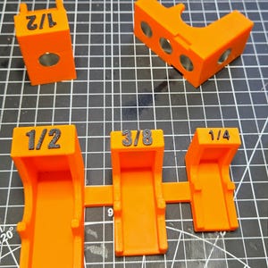 May include: A set of bright orange 3D-printed woodworking jigs. The pieces feature black embossed labels reading 1/2, 3/8, and 1/4. Some components include embedded circular metal magnets, while others have open-ended U-shaped channels for alignment.