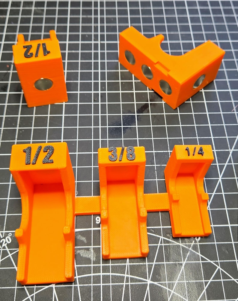 May include: A set of bright orange 3D-printed woodworking jigs. The pieces feature black embossed labels reading 1/2, 3/8, and 1/4. Some components include embedded circular metal magnets, while others have open-ended U-shaped channels for alignment.