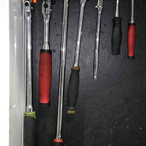 May include: A set of seven chrome-plated metal ratchets with various handle colors including black, red, and green, organized on orange and black plastic holders labeled 1/2, 3/8, and 1/4, mounted vertically inside a tool drawer.