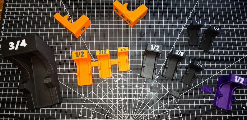 May include: A collection of 3D-printed plastic corner clamps in orange, black, and purple. The clamps are labeled with sizes 3/4, 1/2, 3/8, and 1/4. These L-shaped tools feature distinct markings and varying sizes for woodworking alignment.
