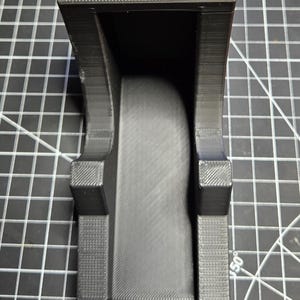 May include: A black 3D-printed plastic holder featuring a prominent white 3/4 label on the top front face. The object has a U-shaped channel design with textured side supports and a smooth central base, designed for organizing or mounting items.