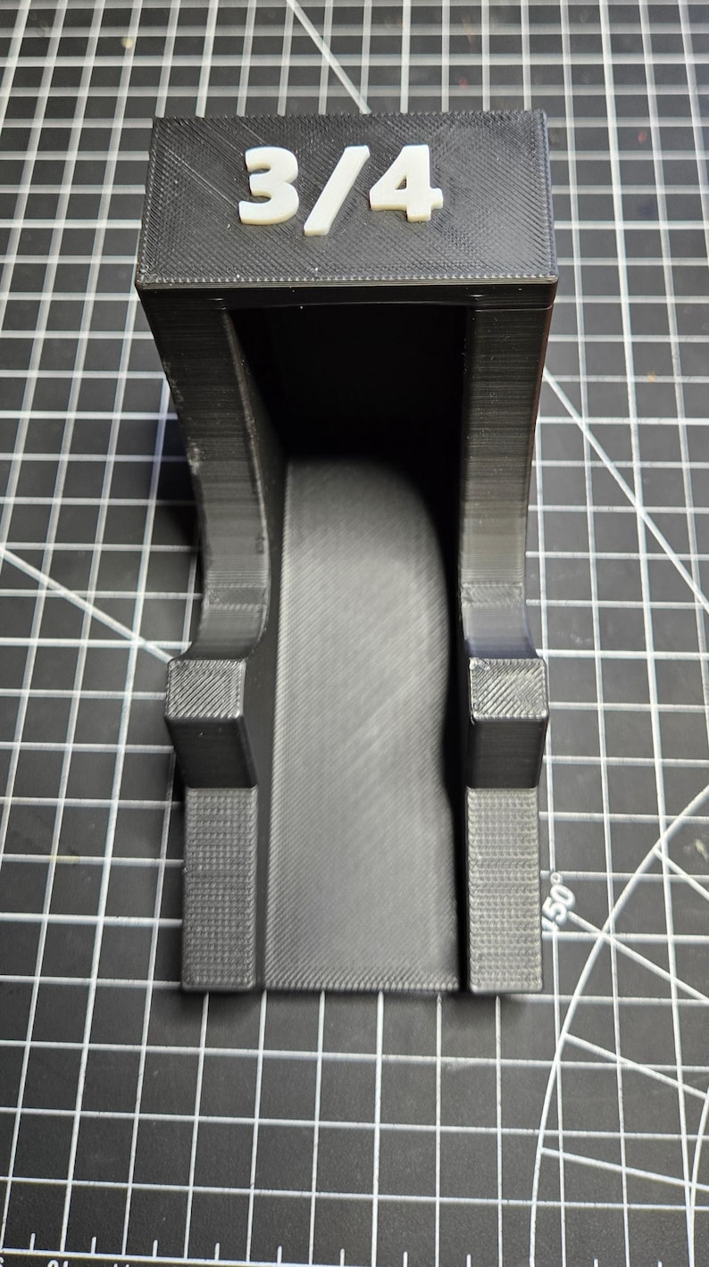 May include: A black 3D-printed plastic holder featuring a prominent white 3/4 label on the top front face. The object has a U-shaped channel design with textured side supports and a smooth central base, designed for organizing or mounting items.