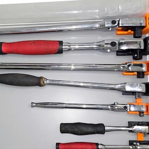 May include: A collection of chrome-plated metal ratchets with various handle colors including red and black, organized in a drawer using orange and black plastic holders labeled 1/2, 3/8, and 1/4 to indicate their respective drive sizes.