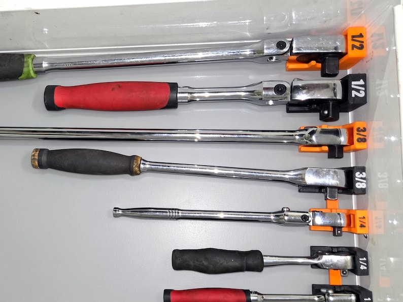 May include: A collection of chrome-plated metal ratchets with various handle colors including red and black, organized in a drawer using orange and black plastic holders labeled 1/2, 3/8, and 1/4 to indicate their respective drive sizes.