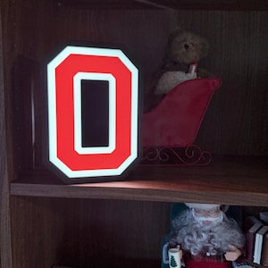 Puede incluir: Una letra "O" roja y blanca iluminada sobre una estantería de madera. Un oso de peluche en un trineo rojo y una figura de Papá Noel también están en la estantería. Un pequeño letrero dice "Santa's on His Way".