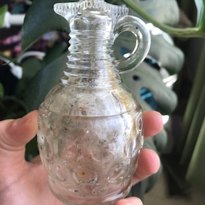 May include: A clear glass cruet with a handle and a decorative design. The cruet has a small spout and a round body with a pattern of circles. The cruet is held in a hand.