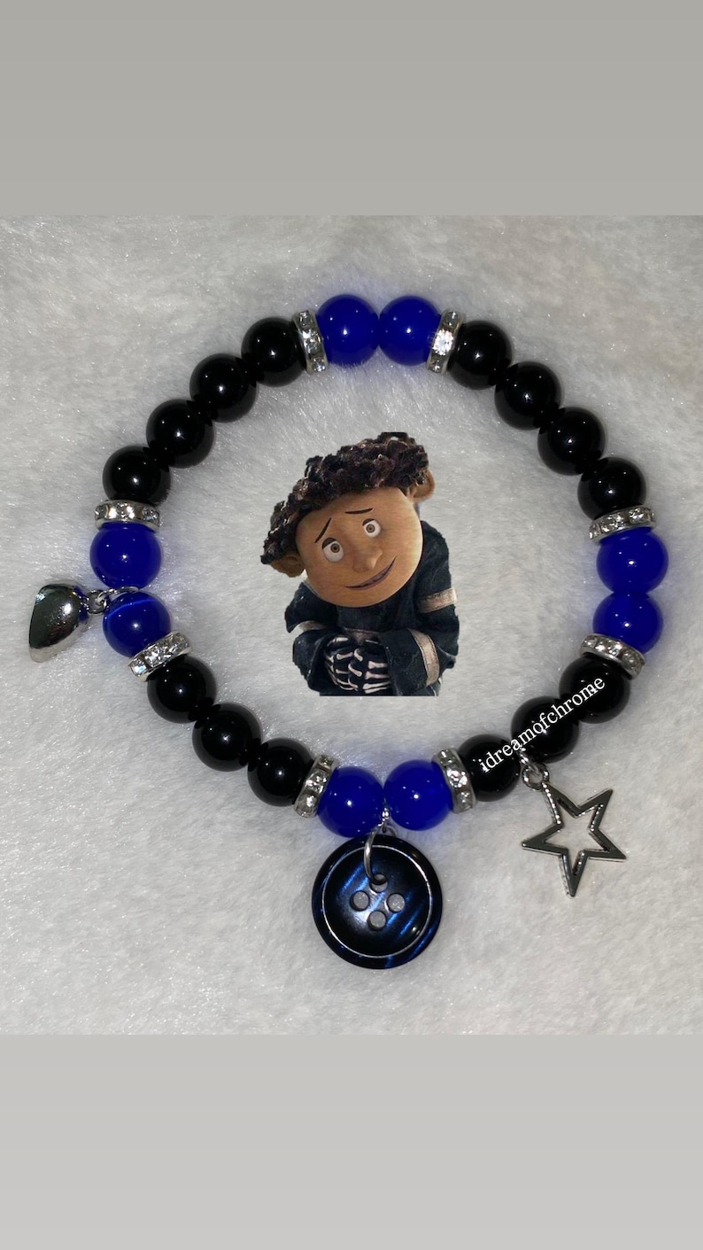 K&ouml;nnte beinhalten: Ein Armband mit schwarzen und blauen Perlen, mit Strass-Abstandshaltern. Es hat Charms, darunter ein Stern, ein Knopf und ein silbernes Herz. Ein Cartoon-Bild ist ebenfalls enthalten. Der Text "idreamofchrome" ist sichtbar.