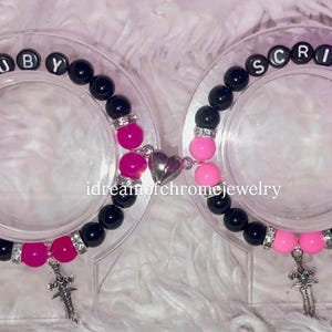 May include: Two matching bracelets with black and pink beads, silver accents, and charms. One bracelet spells "RUBY" and the other spells "SCRIM" with black letter beads. A heart-shaped charm connects the bracelets.