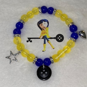 K&ouml;nnte beinhalten: Ein Armband mit gelben und blauen Perlen, silbernen Akzenten und Anh&auml;ngern. Das Armband hat einen Sternanh&auml;nger, einen Herzanh&auml;nger und einen schwarzen Knopfanh&auml;nger. Im Hintergrund befindet sich ein Cartoon-Bild.