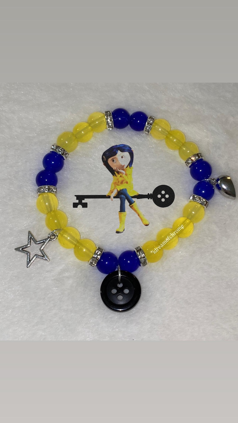 K&ouml;nnte beinhalten: Ein Armband mit gelben und blauen Perlen, silbernen Akzenten und Anh&auml;ngern. Das Armband hat einen Sternanh&auml;nger, einen Herzanh&auml;nger und einen schwarzen Knopfanh&auml;nger. Im Hintergrund befindet sich ein Cartoon-Bild.