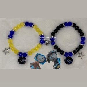 K&ouml;nnte beinhalten: Zwei Armb&auml;nder mit Perlen. Eines ist gelb und blau, das andere schwarz und blau. Jedes Armband hat einen Knopf, einen Stern und einen Herzanh&auml;nger. Die Armb&auml;nder sind vom Film "Coraline" inspiriert.