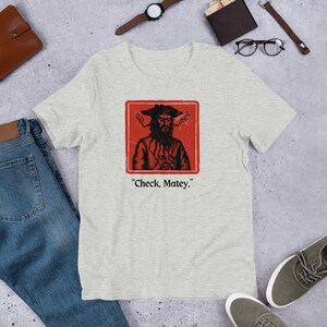 May include: A heather grey t-shirt with a red square featuring a cartoon pirate with a black beard and a black hat. The text "Check, Matey." is printed below the square.