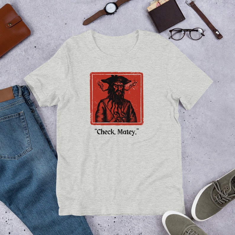 May include: A heather grey t-shirt with a red square featuring a cartoon pirate with a black beard and a black hat. The text "Check, Matey." is printed below the square.