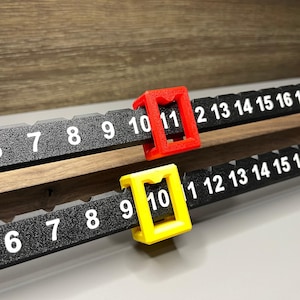 May include: Two black measuring rulers with white numerals and red and yellow markers. The rulers are marked with numbers from 6 to 16. The red marker is on the number 11, and the yellow marker is on the number 10. The rulers are made of plastic.