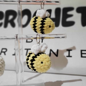 May include: Two crocheted bumblebees with black and yellow stripes and white fuzzy wings hanging from silver keychains on a clear acrylic display stand.