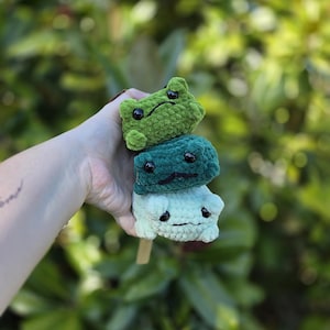 May include: A stack of three small, plush, crocheted frog figures in varying shades of green. The top frog is light green, the middle is dark forest green, and the bottom is a pale mint green. Each frog features black plastic eyes and stitched mouths.