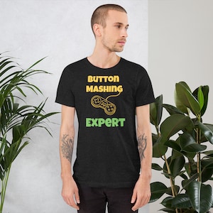 May include: A black t-shirt with a yellow and green graphic of a video game controller. The text "Button Mashing Expert" is printed on the shirt.