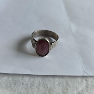 Puede incluir: Un anillo de plata con una piedra preciosa ovalada de color púrpura rojizo. La banda tiene un diseño texturizado. El anillo está sobre un fondo blanco.