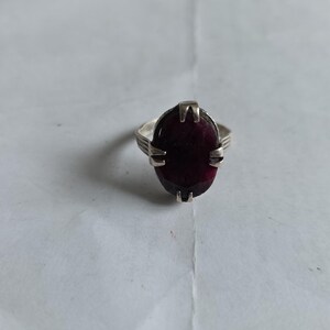 Puede incluir: Un anillo de plata con una gran gema roja oscura de corte ovalado. El anillo tiene una banda sencilla con un diseño decorativo en los laterales, y la gema está sujeta por cuatro garras.