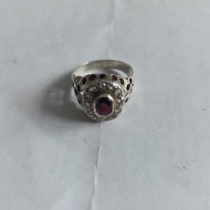 Puede incluir: Anillo de plata con una piedra central ovalada de color rojo oscuro, rodeada de piedras blancas más pequeñas y una banda decorativa. La banda del anillo tiene un diseño ornamentado con detalles en rojo oscuro.