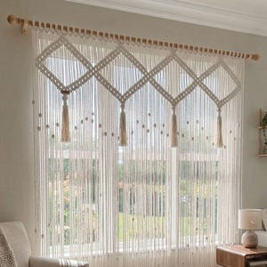 May include: Cream-colored macrame window curtain with a geometric diamond pattern and hanging tassels. The curtain is suspended from a wooden rod, creating a bohemian and textured aesthetic. The curtain is in front of a window.