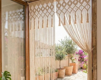 May include: A macrame curtain hangs in a doorway, with a diamond pattern at the top and tassels. The curtain is a natural beige color, and the doorway is framed in wood. A woven basket with a plant sits on the floor.