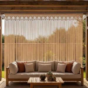 May include: Outdoor patio set with a wooden frame sofa, beige cushions, and brown accent pillows. A matching wooden coffee table sits in front of the sofa. A macrame curtain hangs from a wooden pergola, creating a cozy outdoor living space.