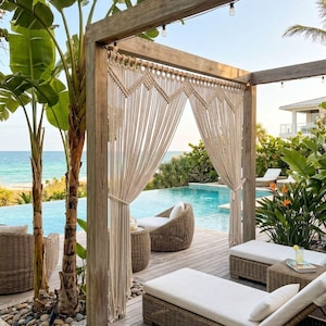 Pode incluir: Uma estrutura de pérgola de madeira com cortinas de macramé em tom off-white com um padrão geométrico de nós, suspensas numa viga horizontal. As cortinas pendem verticalmente, emoldurando uma piscina com mobiliário de vime e plantas.