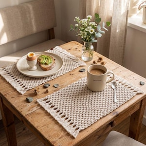 May include: A rustic wooden table set for breakfast. Two woven placemats with fringe detail hold a plate with avocado toast and a soft-boiled egg, and a mug of coffee. A small vase of flowers adds a touch of freshness.