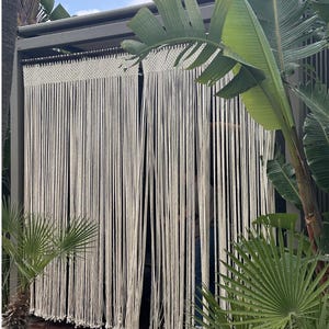 May include: A cream-colored macrame curtain featuring a woven top header with long, hanging vertical cords that terminate in small, knotted ends. The curtain is suspended from a dark horizontal rod, creating a textured, linear partition.